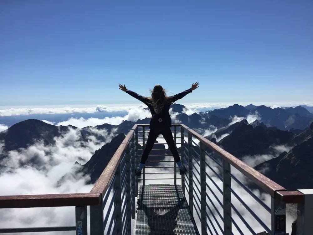 woman standing on a guardrail overlooking clouds and mountaintops.
